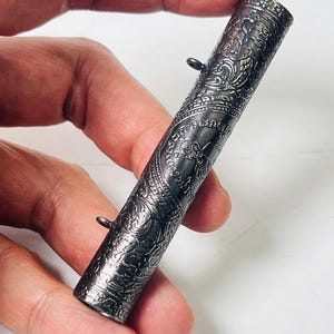 Puede incluir: Un recipiente cilíndrico de metal plateado con intrincados patrones grabados. El recipiente tiene un pequeño lazo a cada lado y parece ser un artículo decorativo o ceremonial. La superficie está detallada con motivos florales y figurativos.