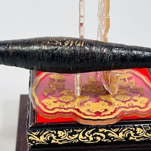 May include: A dark, cylindrical object with a textured surface, possibly made of wood or a similar material. It is suspended above a decorative base with a red and gold design. A small, round, bronze-colored medallion hangs from the object.