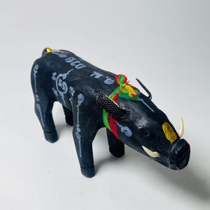 Può includere: Statuetta in legno intagliato di un cinghiale nero con zanne bianche e dettagli dorati. Il cinghiale presenta dettagli dipinti e scritte. Un nastro colorato è legato attorno a un orecchio.