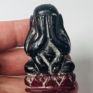May include: A small, dark sculpture of a figure covering its eyes with its hands. The figure is seated on a red base with a carved design. The sculpture is held in a person's hand.