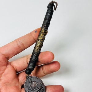 May include: A dark brown pendant with a detailed metallic medallion. The pendant has a cylindrical handle with gold accents and a woven cord. The medallion features an engraved image of a seated figure and text.