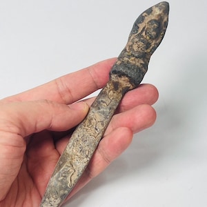 May include: A weathered letter opener with a stone-like appearance. The handle and blade display a mottled pattern of grey, brown, and beige. The opener is held in a hand against a white backdrop.