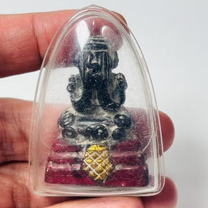 May include: A small, black figurine of a deity encased in a clear, arched plastic case. The figurine sits on a red base with a gold-colored, textured design. The case is held in a person's hand.