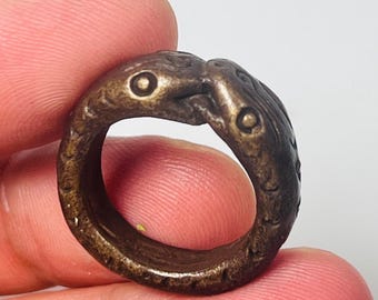Könnte beinhalten: Ein bronzefarbener Ring mit zwei vogelartigen Figuren, die sich gegenüberstehen. Der Ring hat eine strukturierte Oberfläche und detaillierte Schnitzereien mit sichtbaren Augen und Schnäbeln. Ein einzigartiges Schmuckstück.
