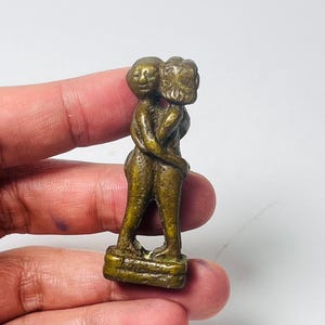 Puede incluir: Una pequeña escultura de color bronce que representa dos figuras abrazándose. Las figuras están estilizadas con rasgos redondeados y están de pie sobre una base rectangular. La escultura tiene un aspecto vintage.