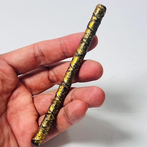 Puede incluir: Una varita decorativa dorada y negra. La varita es cilíndrica con una superficie texturizada. La pintura dorada se aplica en un patrón de mosaico sobre una base negra. La varita se sostiene en una mano.
