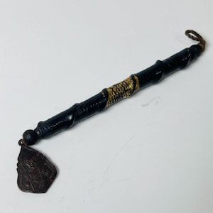 May include: A black, cylindrical object with a gold-colored band and a brown pendant. The pendant has a detailed design. The object has a twisted, textured appearance and a loop at the end.