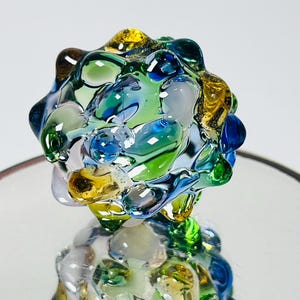 May include: A colorful, abstract glass sculpture. The piece features a rounded, textured form composed of fused glass in shades of blue, green, yellow, and clear. The glass has a glossy finish, reflecting light and creating a dynamic visual effect.