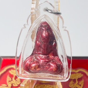 Op de afbeelding: Een rode en gouden hanger met een Boeddha-figuur, ingekapseld in een heldere, beschermende behuizing. De hanger is opgehangen aan een zilverkleurige ring. De achtergrond is een rood en gouden decoratief oppervlak.