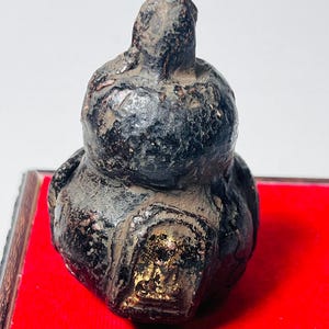 May include: A small, dark-coloured, weathered sculpture with a bulbous base and a rounded top. The sculpture has a rough texture and appears to be made of a dark material with gold accents. It sits on a red surface.