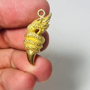 Puede incluir: Colgante dorado con un diseño detallado de serpiente enrollada y un colmillo prominente. El colgante tiene un ojal para una cadena y está sujeto en la mano de una persona.