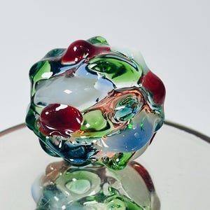 May include: A colorful, abstract glass sculpture with a rounded shape. The sculpture features a mix of colors, including red, green, blue, and white. The glass has a glossy, reflective surface, and the piece is displayed on a reflective surface.