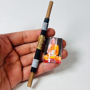 May include: A wooden stick with black and gold accents, and a small picture of a monk in orange robes. The stick has a wrapped design with black thread, gold glitter, and gray tape.