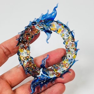 May include: A handcrafted glass sculpture in the shape of a dragon or serpent, with a circular form. The sculpture features vibrant colors, including blue, yellow, red, and white, and is held in a hand against a white background.
