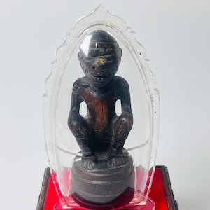 Puede incluir: Una figura de madera marrón oscuro de una figura en cuclillas, encerrada en una pantalla de plástico transparente. La figura está montada sobre una base oscura, colocada sobre una plataforma roja. La figura tiene una cara detallada y está encerrada en una carcasa transparente.