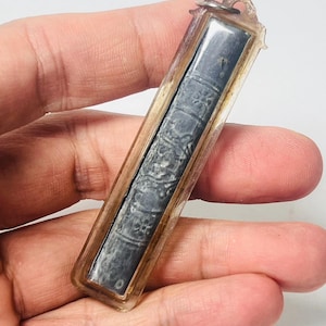 May include: A rectangular, dark gray amulet encased in a clear, rectangular plastic frame. The amulet has intricate, embossed patterns and is held in a hand against a white background. The frame has a small loop at the top.