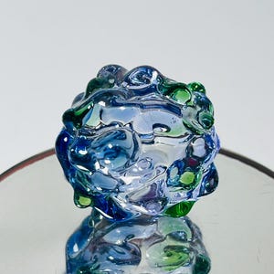 May include: A decorative glass sculpture with a rounded, organic shape. The piece features a blend of blue and green hues, with a glossy, textured surface. The sculpture is displayed on a reflective surface, enhancing its visual appeal.
