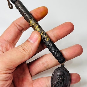 May include: A dark brown pendant with a detailed image of a person in a frame, attached to a cylindrical, textured, dark brown object with gold accents. The pendant is connected to a loop of brown string.