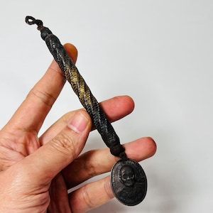 May include: A dark, twisted metal pendant with a skull medallion. The pendant has a braided design with gold accents. The medallion features a detailed skull image. The pendant is held by a hand against a white background.