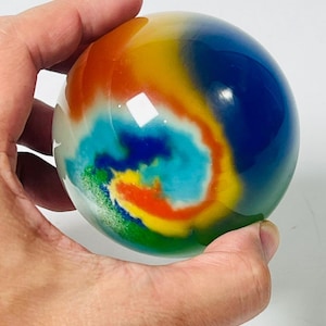 Op de afbeelding: Een gladde, bolvormige glazen presse-papier met een levendig, wervelend patroon van kleuren, waaronder diepblauw, feloranje, geel, turkoois en groen. Het oppervlak is gepolijst en reflecterend, met een gemarmerd, veelkleurig ontwerp.