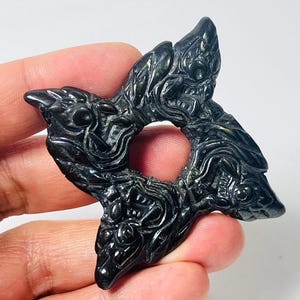 Könnte beinhalten: Ein schwarzer, sternförmiger Anhänger mit aufwendigen, geschnitzten Details. Der Anhänger hat ein zentrales Loch und spitze Kanten, gehalten in einer Hand vor weißem Hintergrund. Das Design zeigt wirbelnde Muster und stilisierte Figuren.