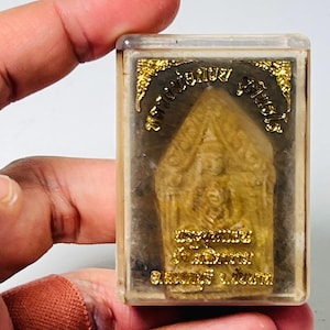 May include: A rectangular clear plastic protective case containing a small, tan-colored, pointed amulet with embossed Thai script and gold-colored decorative accents at the top corners. The amulet features a central figure in relief.