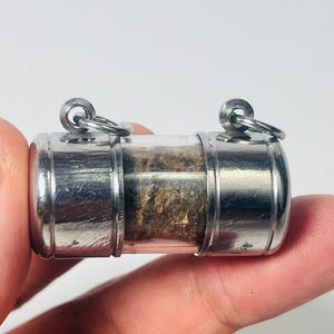 Puede incluir: Un pequeño amuleto cilíndrico encerrado en vidrio transparente con tapas y bucles de metal plateado. El interior contiene un material marrón y texturizado. El amuleto está sostenido por los dedos de una persona sobre un fondo blanco.