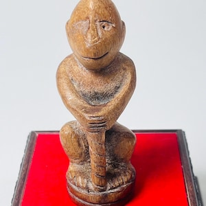 Puede incluir: Figura de mono de madera tallada, con una expresión sonriente, sentado con las manos juntas. El mono es de color marrón claro y se asienta sobre una base roja con un marco decorativo negro y dorado. Un artículo decorativo único.