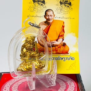 May include: A gold-colored Thai amulet featuring a deity figure, encased in a clear acrylic protective shell with a metal loop. Behind the amulet is a yellow card displaying a photograph of a monk in orange robes and traditional Thai script.