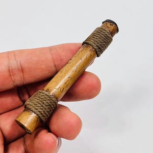 May include: A cylindrical, light brown wooden object with dark brown twine wrapped around both ends. The object has etched markings and a dark cap on one end. The item is held in a hand against a white background.