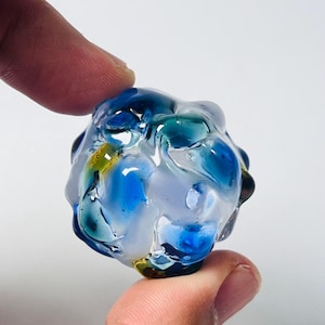 May include: A colorful, abstract glass bead with a rounded shape. The bead features swirls of blue, teal, yellow, and white, creating a unique, artistic design. The glass appears translucent, allowing light to pass through. The bead is held between two fingers.