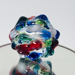 May include: A colorful glass bottle stopper with a rounded, textured design. The stopper features a mix of red, blue, green, and white glass, creating a vibrant and artistic appearance. The stopper is designed to fit on a bottle.