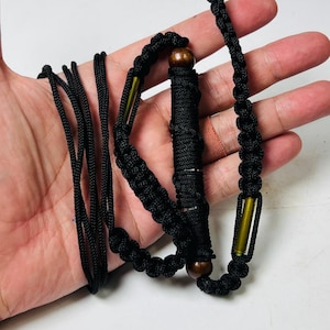 May include: A black paracord lanyard featuring a central cylindrical section wrapped in black cord, accented with two dark brown wooden beads and two metallic olive-green cylindrical spacers, finished with a braided cord strap and long loose cord ends.