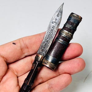 May include: A small, pointed metal blade featuring intricate dark engravings and script, attached to a dark, textured handle with metallic accents. Beside the blade sits a matching cylindrical sheath with dark, patterned casing and metallic bands.