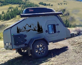 May include: A small, gray teardrop camper trailer with black mountain and tree decals, parked on a dirt road. The trailer has a black storage box on top and a small window. Mountains and a blue sky are in the background.