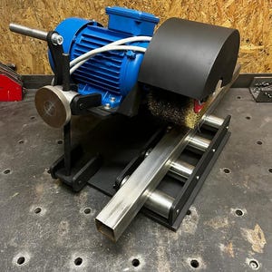 Puede incluir: Un motor eléctrico azul con una cubierta protectora negra y un accesorio de cepillo metálico. La máquina está montada sobre una base negra con un sistema de rodillos metálicos. Un tubo cuadrado de metal está posicionado para el procesamiento.