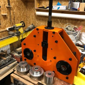 May include: An orange metal fabrication tool with a black handle and several silver metal components. The tool is mounted on a wooden workbench, with additional metal pieces nearby. The background shows a workshop setting.