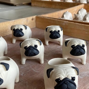 以下が含まれることがあります： セラミック製のパグの植木鉢のコレクション。植木鉢はオフホワイトで、顔、耳、目の周りに黒のアクセントがあります。植木鉢の中には、植えるための開口部があるものもあります。