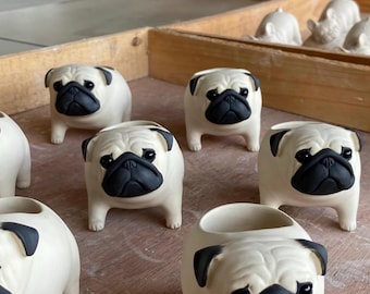 Puede incluir: Una colección de macetas de cerámica con forma de pug. Las macetas son de color blanco roto con detalles negros en la cara, las orejas y alrededor de los ojos. Algunas macetas tienen la parte superior abierta para plantar.