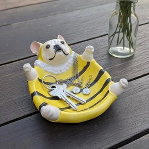 May include: A novelty ceramic bulldog-shaped trinket dish in yellow and black stripes, holding keys, a necklace, and earrings. The bulldog has a white face and pink ears. The dish sits on a dark wooden surface.