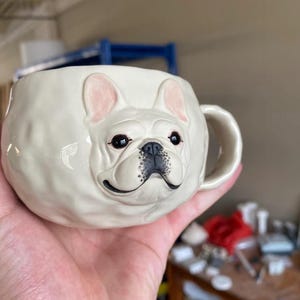 以下が含まれることがあります： フレンチブルドッグの頭の形をした白いセラミックマグ。マグカップは、黒い目、黒い鼻、ピンクの裏地のある耳を備えた詳細な顔を特徴としています。ハンドルはシンプルなループで、マグカップは手で持たれています。