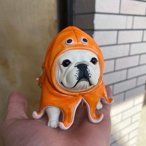 May include: A ceramic figurine of a French Bulldog wearing an orange octopus costume. The dog has a white face with black markings and large, dark eyes. The octopus hood has two large, black eyes and tentacles.