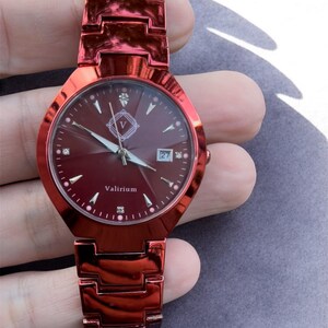 May include: A red wristwatch with a matching strap. The watch face is a deep red with diamond-shaped hour markers and a date display. The brand name "Valirium" is printed on the face. The watch is held in a hand.