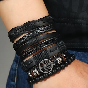 Puede incluir: Una colección de pulseras negras con varios diseños. Las pulseras incluyen cuero trenzado, cuentas y bandas de cuero. Una pulsera presenta un dije plateado del árbol de la vida. Las pulseras están apiladas en una muñeca.
