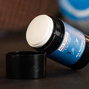 May include: A black and blue sports product with a white top and a detached black cap. The product has a blue label with white text that reads "ALL SPORT Blister Prevention & Anti-Chafe ALL NATURAL".