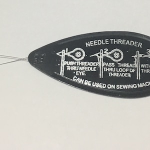 Könnte beinhalten: Schwarzer Plastik-Nähfaden-Einfädler mit einer silbernen Metallschlaufe. Der Einfädler hat bedruckte Anweisungen: "Push threader thru needle eye. Pass thread thru loop of threader. Withdraw threader. Can be used on sewing machines."