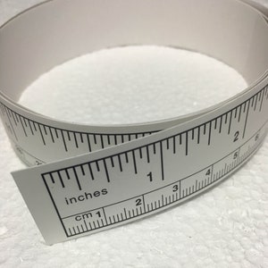 May include: White measuring tape with black markings in inches and centimetres. The tape is rolled up and the markings are visible.