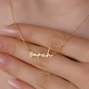 Peut inclure: Un délicat collier en or avec le nom "Sarah" écrit en cursive. Le pendentif est suspendu à une fine chaîne, créant un accessoire personnalisé et élégant. Le collier est présenté sur un fond neutre.