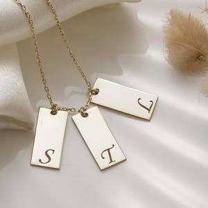 Può includere: Una collana dorata con tre ciondoli rettangolari, ognuno inciso con una lettera: "S", "T" e "J". La catena è delicata e i ciondoli sono posti su uno sfondo bianco e setoso.
