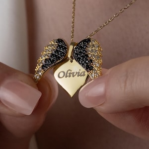 May include: A gold-colored necklace featuring a pendant with the name "Olivia" engraved on a heart-shaped tag. The pendant is flanked by two wing-shaped elements, adorned with black and clear gemstones. The necklace is held by a person.